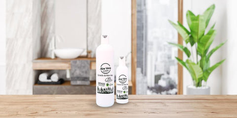Image of two bottles of Aloe Vera Extract product of the brand Aloe Vera Exclusive on a wooden surface. The larger bottle is behind a smaller one, both with a minimalist design. In the background, you can see the interior of a modern bathroom with white curtains, a shelf with towels and a plant, suggesting a calm and relaxing atmosphere.