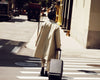 Man in beige overcoat walks down New York street with black suitcase trailing behind