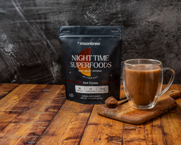 Packet of Moonbrew Nighttime Superfoods Hot Cocoa with glass mug of hot cocoa on wooden surface.