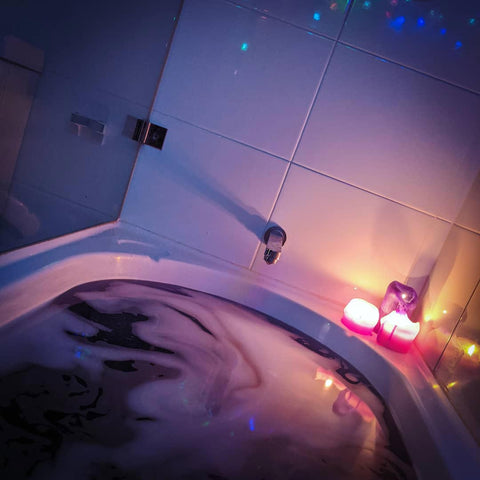 A dark purple, frothy bath, with 3 pink/purple candles on the right corner, and the reflection of coloured fairy lights on the tiles