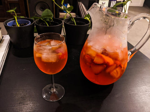 A wine glass and clear jug containing a reddish cocktail with ice cubes and strawberries