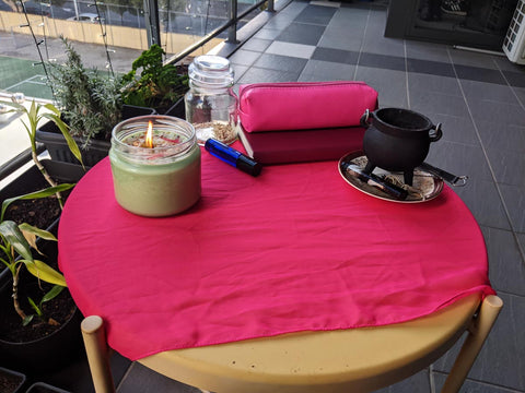 A small round table on a balcony with plants. The table has a pink altar cloth, and contains a green candle, small cauldron, notebook, pencil case, and a jar.