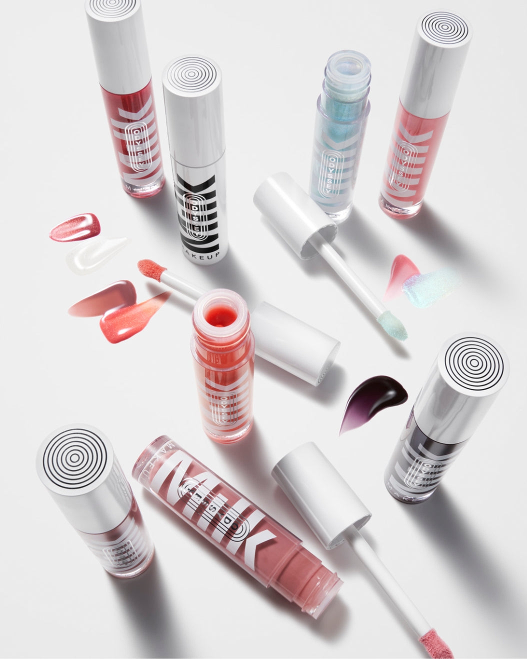 Product shot of every tube of Milk Makeup Odyssey Lip Oil Gloss arranged with their swatches and applicators, on a white background.