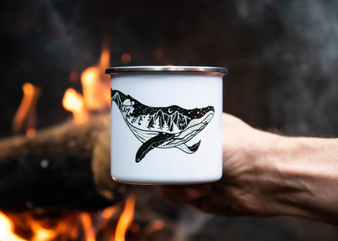 A hand holding an enamel mug with an illustrated whale on it in front of campfire 