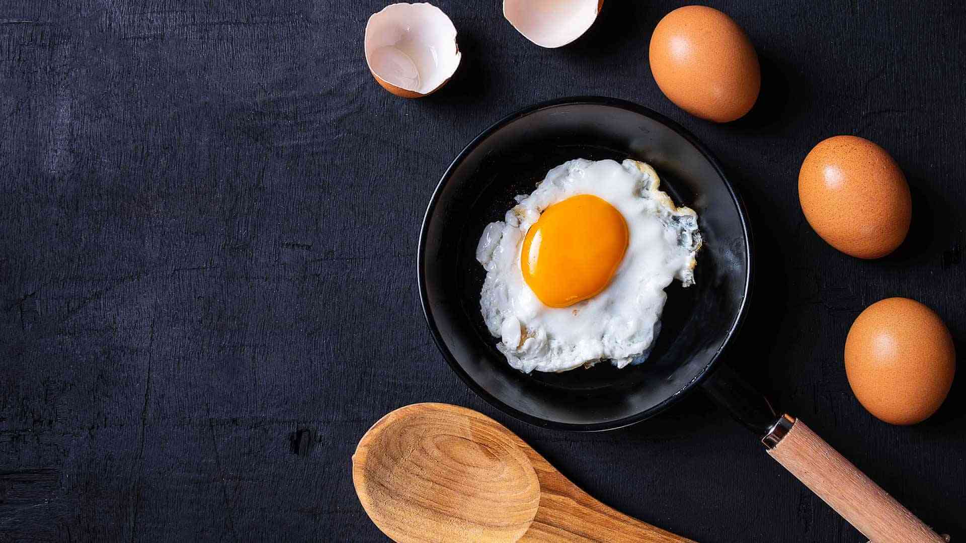 Sunny side up egg on a pan that's surrounded by cracked and uncracked brown eggs and a wooden spatula.