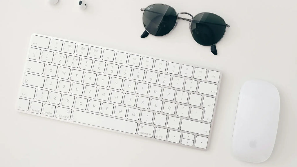 magic mouse next to a keyboard and sunglasses on desk