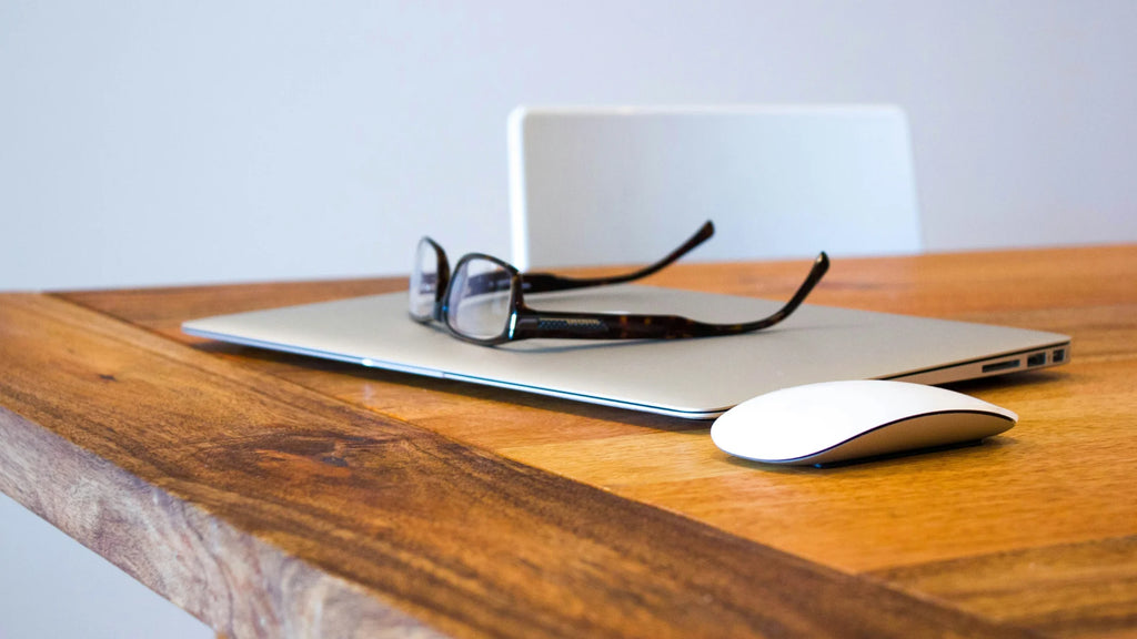 Brown desk with Magic Mouse and Macbook with Glasses on top