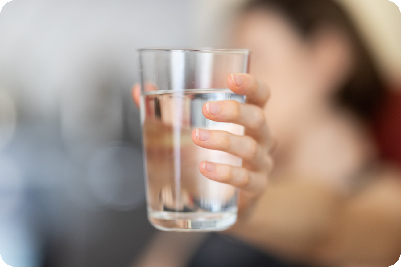 Person hält ein großes Glas klares Wasser in die Kamera, symbolisch für den Tipp, den Tag mit ausreichender Hydratation zu beginnen, als wesentlicher Bestandteil einer erfolgreichen und energiereichen Morgenroutine.