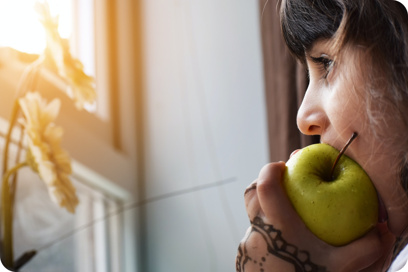 Nachdenkliche Frau schaut aus dem Fenster und hält einen Apfel im Mund, symbolisch für den Nutzen von Äpfeln als energiereiche Snacks, die bei der Linderung von Müdigkeit und Energiemangel helfen können.