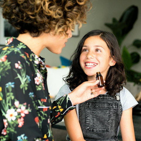 A woman holding a bottle of essential oil, smiling at a young girl in a denim jumper