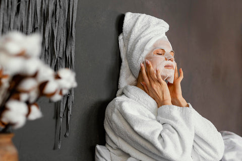A woman in a robe and towel on her head applying a facial mask for a skincare routine