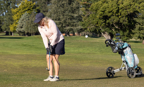 Woman wearing petal collared golf top, navy golf skort chipping the ball towards the camera