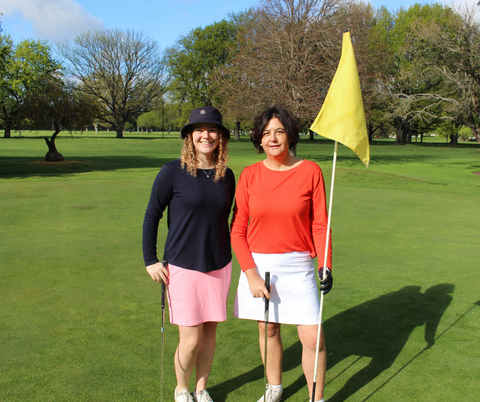 Harriet and Liz in golf skorts standing on golf course with a yellow flag