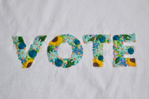 Hand embroidered floral block letters in blues and yellows with orange and green accents. The block letters spell out VOTE on a white tshirt. The flowers are sunflowers, daisies, and roses with green leaves and little french knots.