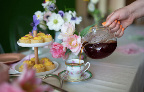 A person's hand pouring black tea from a pot into a small cup, beside a tea tower of lemon curd cake bites.