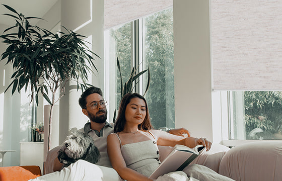 A couple is reading on the sofa, and linen stone roller shades are installed on the window next to them.