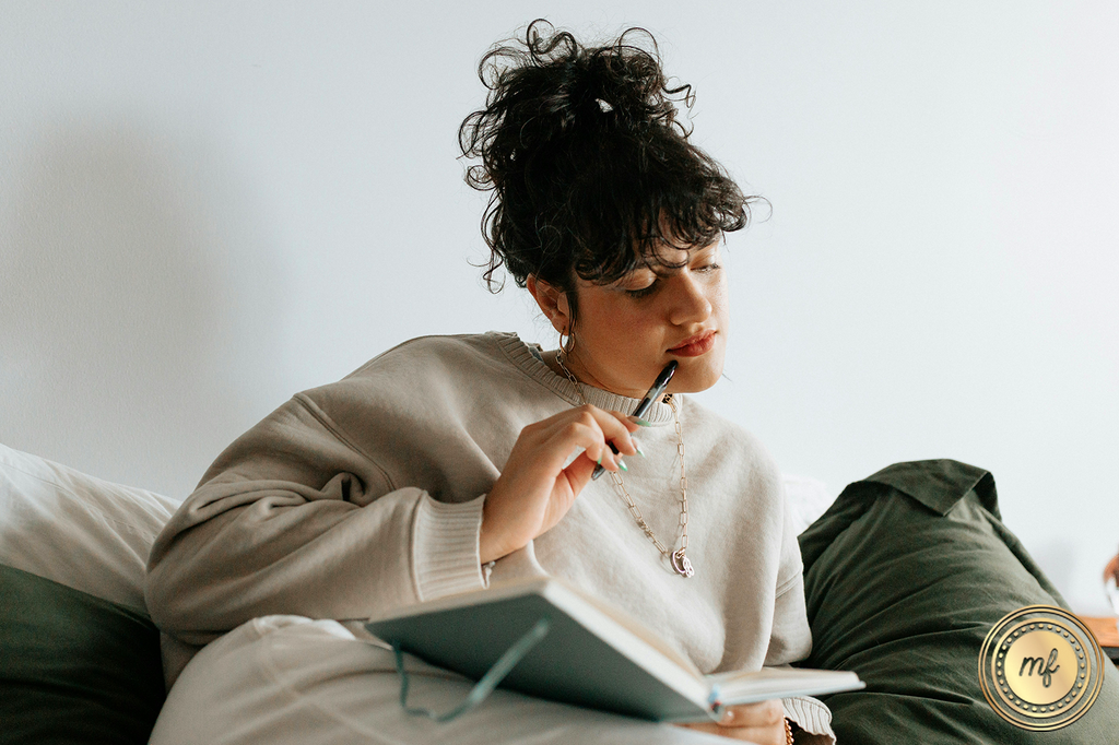 A smiling woman sits, holds a pen, and focuses on reading a book