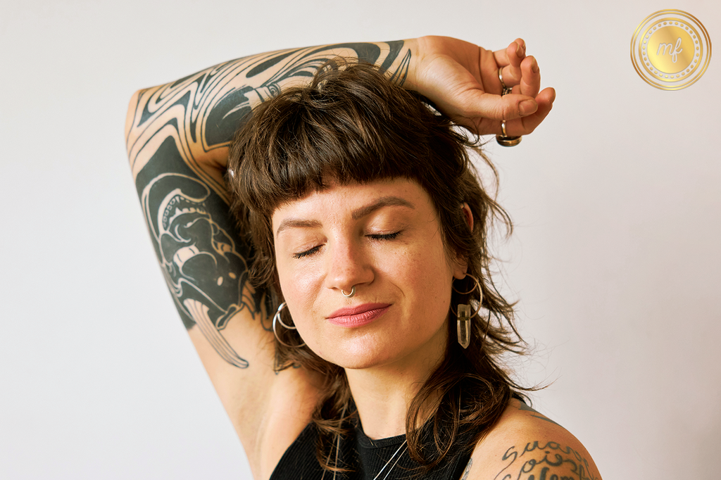 A smiling woman, hand resting on her head, eyes closed, showcasing a tattoo on her hand