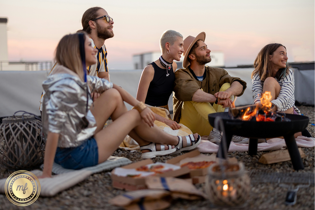 Silhouetted figures sit on rooftop at sunset, gathered around fire pit, enjoying evening ambiance