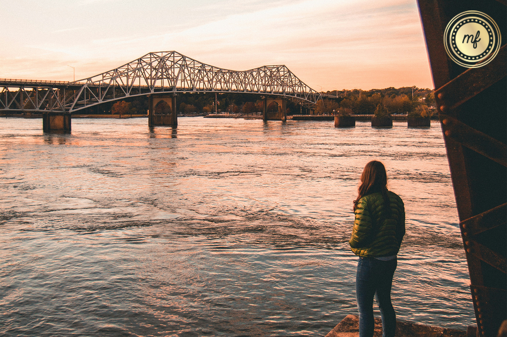 A woman, standing by the riverside at sunset, looks towards a bridge spanning the river