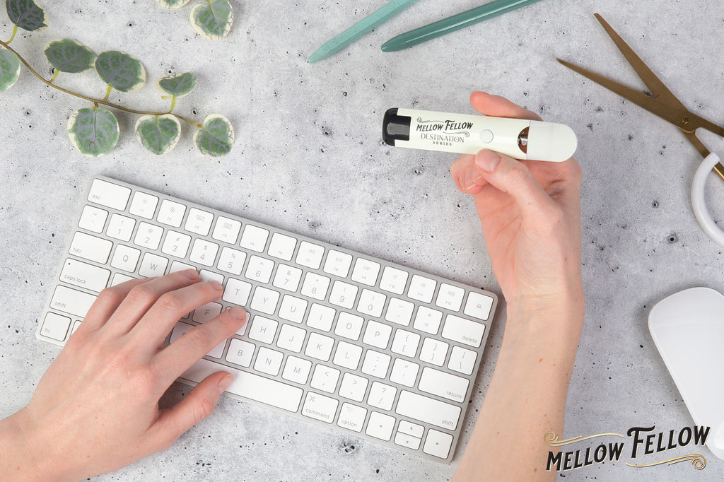 A hand types on a white keyboard while the other holds a "Mellow Fellow" DESTINATION series vape device, surrounded by a green pencil, scissors, mouse, and a leafy sprig on a gray surface