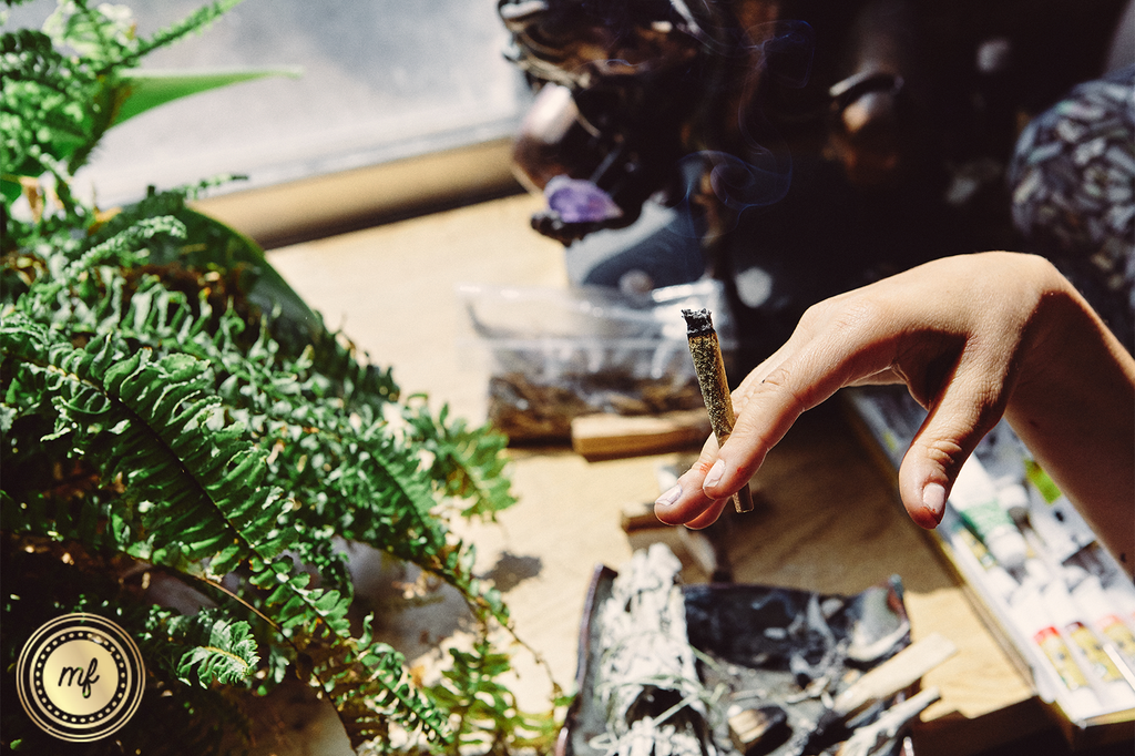 Hand holding a joint on a table with plants and an ashtray.