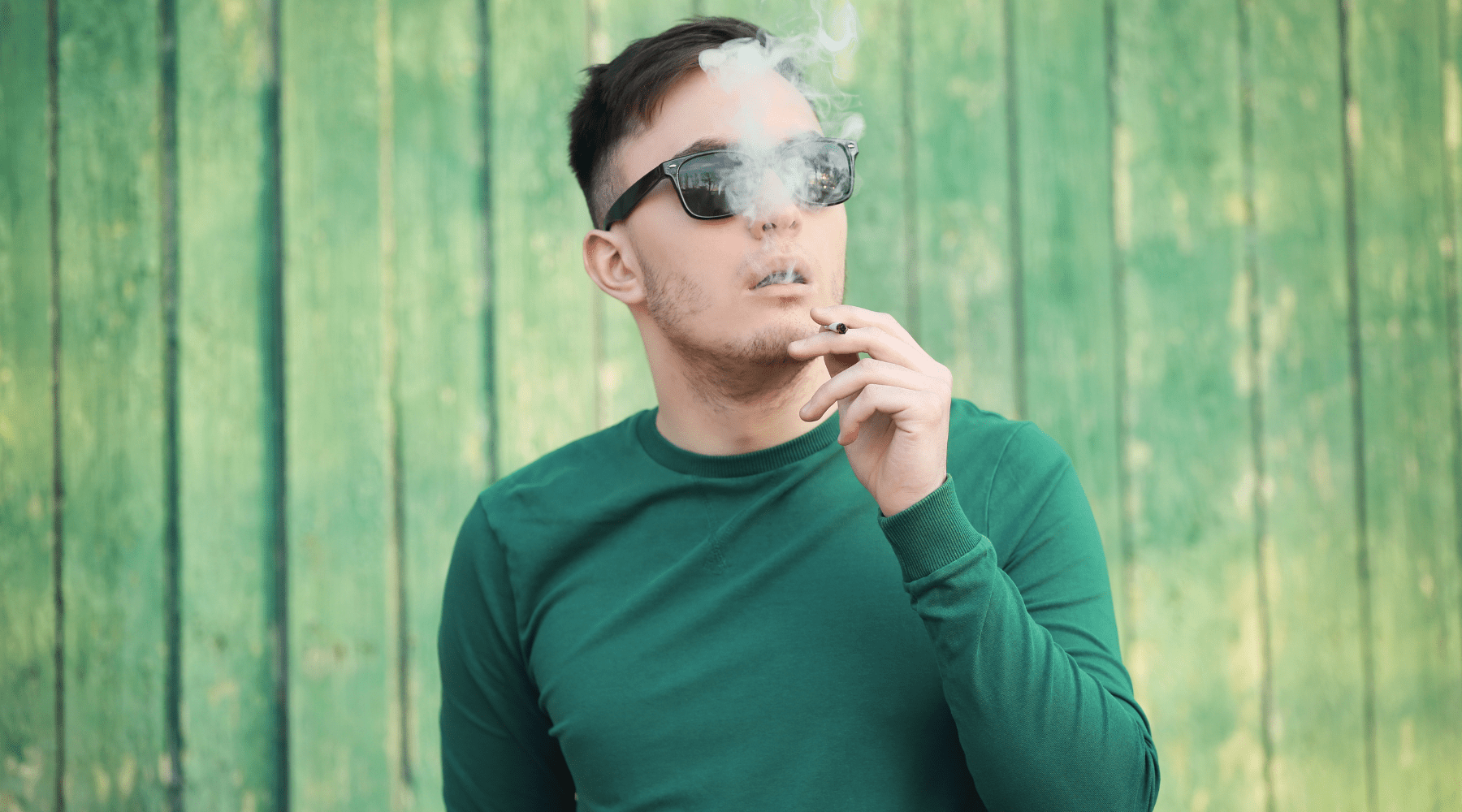 a photo of a man in a green sweater smoking a joint rich in delta-9 THC in front of a green fence