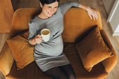 A smiling pregnant woman relaxing on a mustard-colored sofa, holding a warm cup of tea.