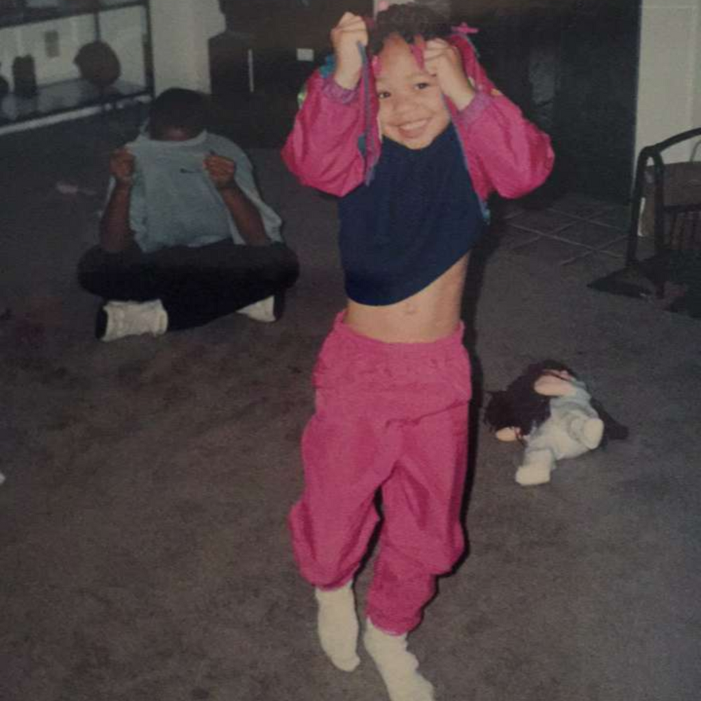 Denine as a child wearing a pink track suit trying to hide from the camera.