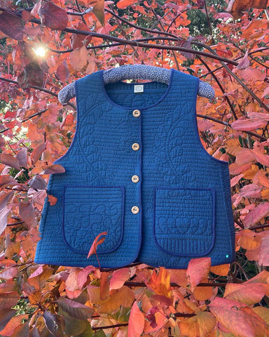 Blue quilted waistcoat/gilet/body warmer/vest with wooden buttons and patch pockets hanging among some red autumn leaves