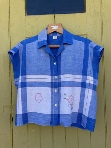Boxy shirt made from a blue and white checked tablecloth with floral embroidery. Hanging on a rather dirty mustard yellow garage door