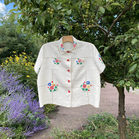 Cream-coloured blouse with floral appliqué and red buttons, made from a vintage tablecloth. Hanging from a tree in a park