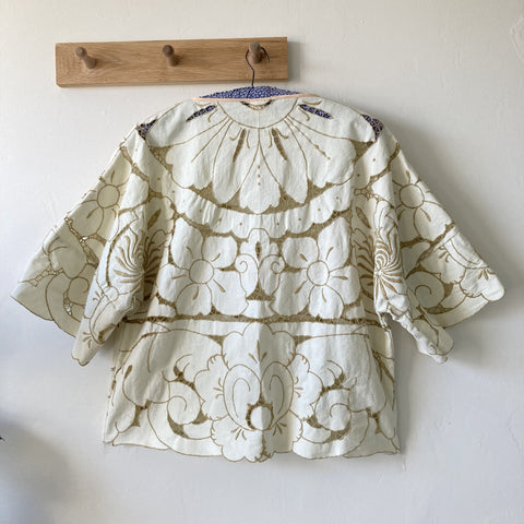 Back view of a boxy, short-sleeved top made from a cream-coloured cutwork tablecloth, hanging on some wooden hooks