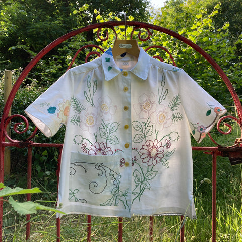 A shirt made of recycled embroidered tablecloths. It is mostly white with colourful stitched flowers. It's hanging on a red-painted garden gate