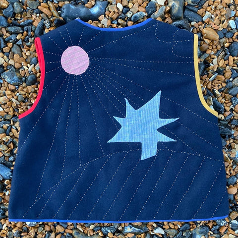 Hand quilted vest/gilet/body warmer/waistcoat with a landscape, tree and sunrays appliqué and stitched onto the back/ Multicoloured binding. Photographed on a pebbly beach