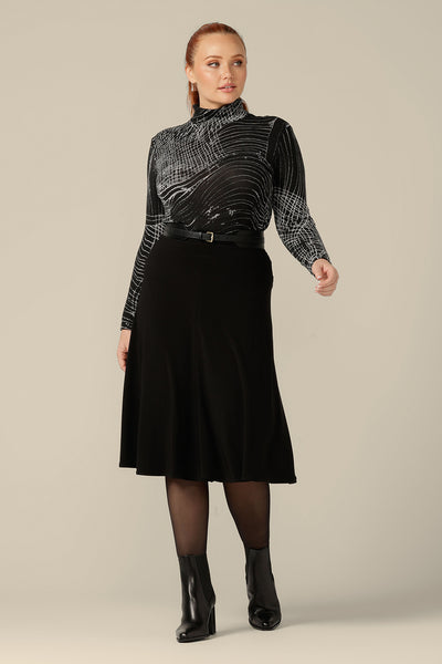 A curvy woman wears a long sleeve, woolly knit turtleneck top, size 12, in an abstract black and white pattern. A warm winter top, this women's poloneck top is worn with a flared, knee length black skirt and faux leather belt. 