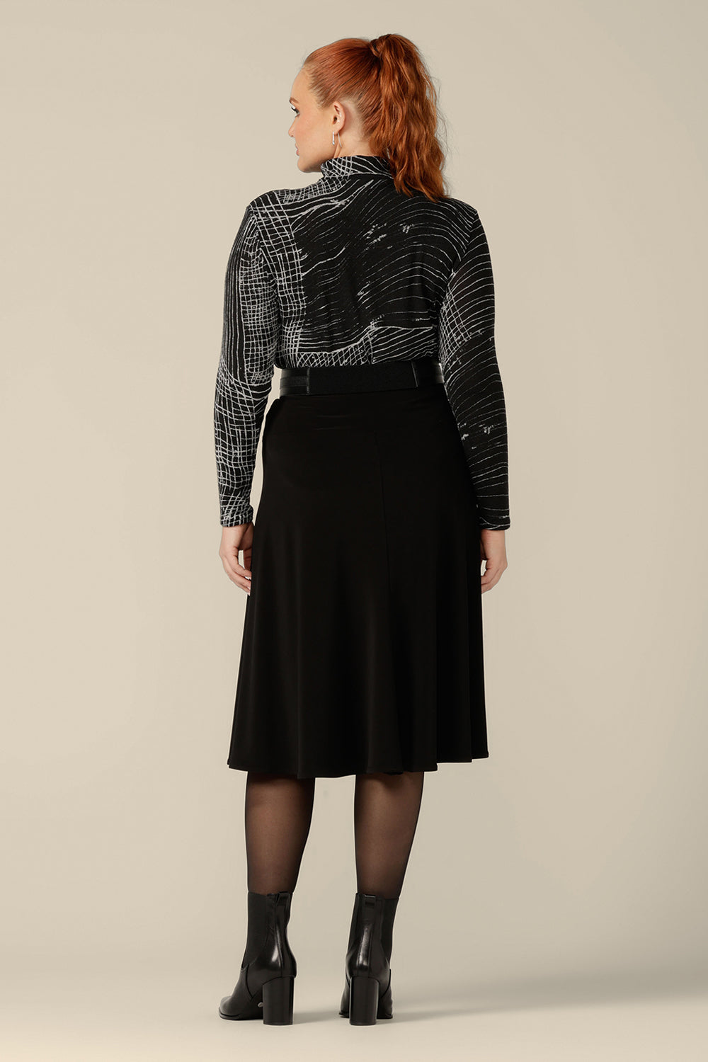 Back view of a curvy woman wearing a long sleeve, woolly knit turtleneck top, size 12, in an abstract black and white pattern. A warm winter top, this women's poloneck top is worn with a flared, knee length black skirt and faux leather belt. 