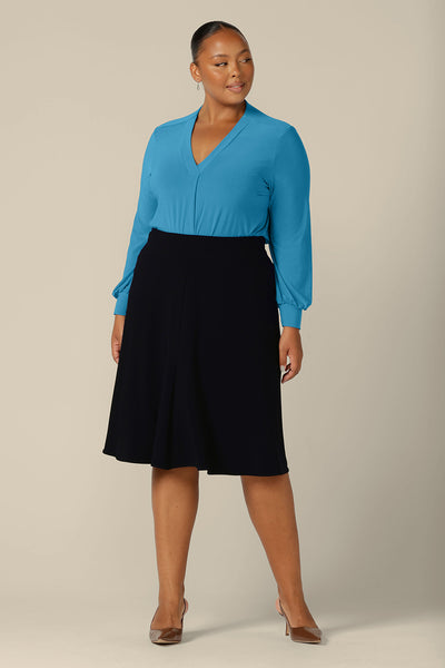 A fuller figure woman wears a V-neck top in size 18 with long bishop sleeves in Opal blue stretch jersey. Made in Australia by Australian and New Zealand women's clothing label, L&F, this workwear top is worn with a knee-length navy skirt for a complete workwear outfit. 