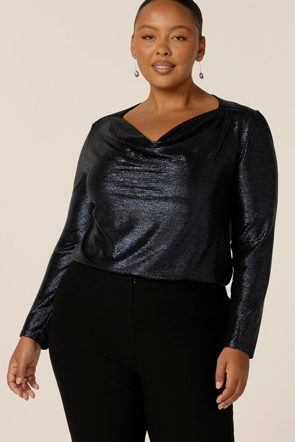 A size 18 woman wears a long sleeve occasionwear top with cowl neck in shimmering midnight blue jersey fabric. A great evening wear top for fuller figures, this long sleeve top wears well for special events and cocktail parties. Made in Australia, this semi-formal top sparkles in sizes 8 to 24 .