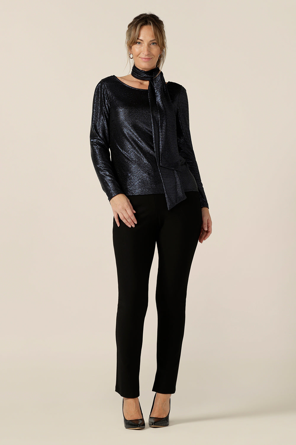 A size 10, 40-plus woman wears a shimmering midnight blue sparkly top with high scoop neck and long sleeves. Worn with slim leg, black evening pants, and shimmering blue neck tie, this is a simple and easy-to-wear, comfortable eveningwear top. Made in Australia in sizes 8 to 24.
