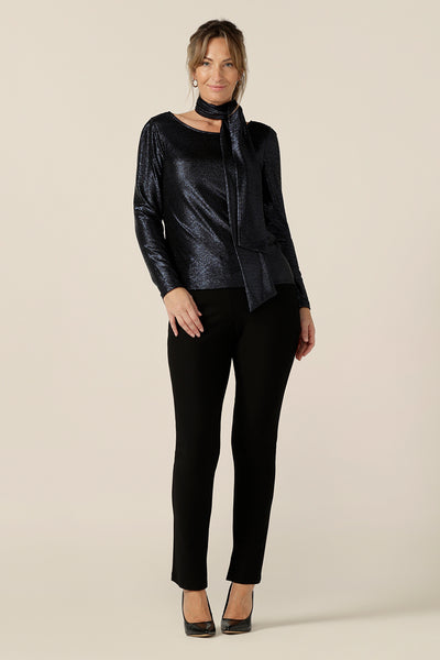 A size 10, 40-plus woman wears a shimmering midnight blue sparkly top with high scoop neck and long sleeves. Worn with slim leg, black evening pants, and shimmering blue neck tie, this is a simple and easy-to-wear, comfortable eveningwear top. Made in Australia in sizes 8 to 24.