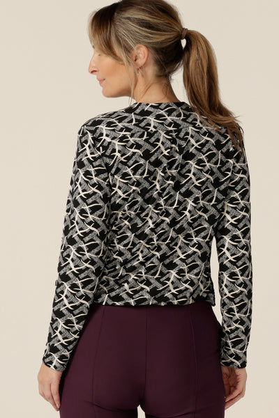 Back view of a size 10 woman wearing a V-neck jersey top with long sleeves and a shirttail hemline. Made in Australia by women's workwear and casual wear brand, L&F, this long sleeve top features a black and white graphic print.