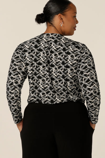 Back view of a size 18, plus size woman wearing a V-neck jersey top with long sleeves and a shirttail hemline. Made in Australia by women's workwear and casual wear brand, L&F, this long sleeve top features a black and white graphic print.