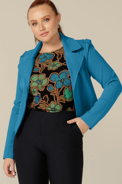 A size 12, curvy woman wears a floral print, boat neck top with long sleeves under an Opal blue soft tailored jacket.  Both are made in Australia by size inclusive clothing label, L&F.