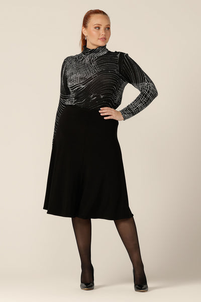 A curvy woman wears a long sleeve, woolly knit turtleneck top, size 12, in an abstract black and white pattern. A warm winter top, this women's poloneck top is worn with a flared, knee length black skirt, both made in Australia.