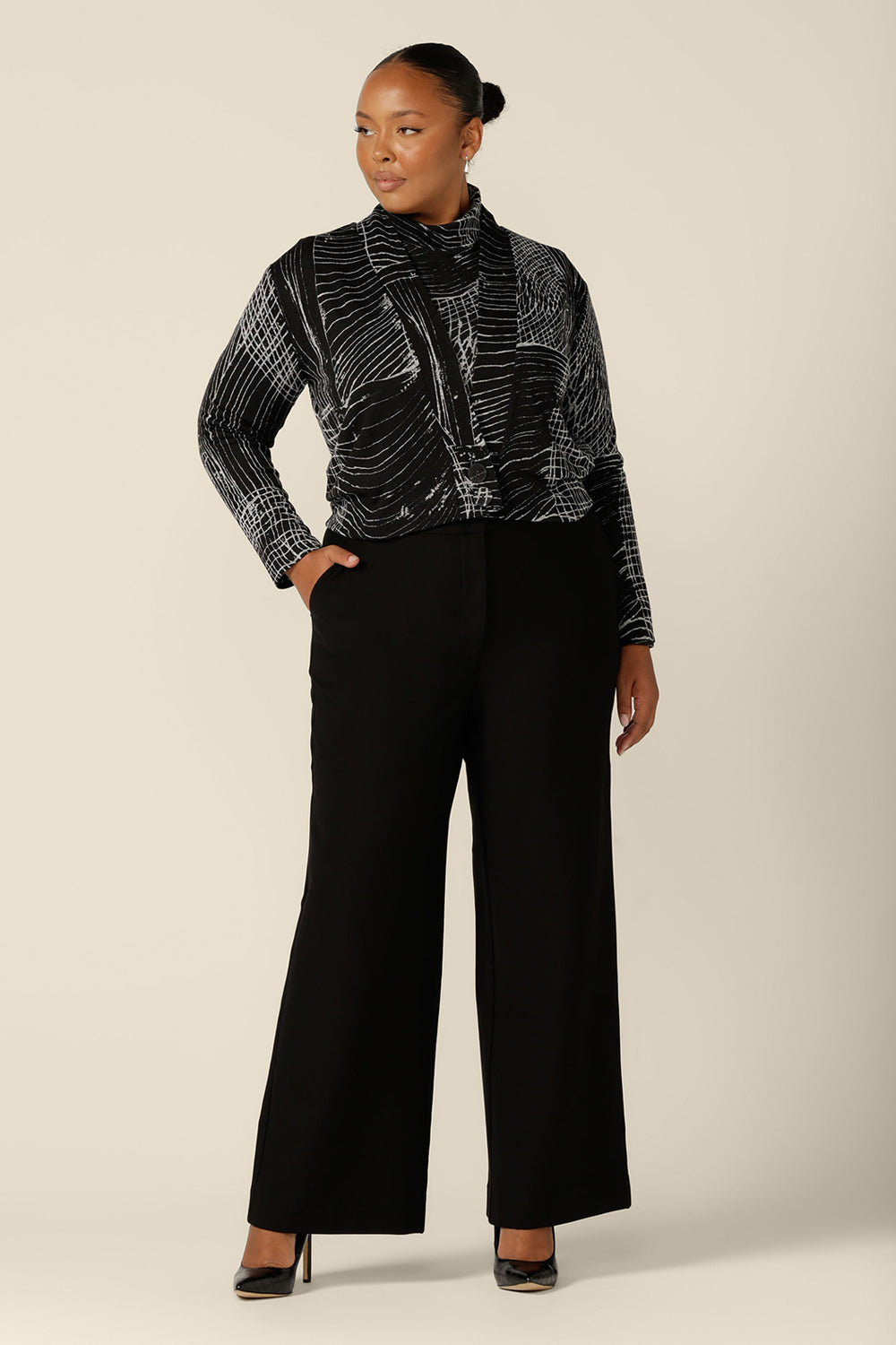 A fuller figure woman wears a long sleeve, woolly knit turtleneck top, size 18, in an abstract black and white pattern. A warm winter top, this women's polo neck top by Australian and New Zealand women's clothing label, L&F is worn with high-waisted, wide leg black pants and woolly knit cardigan.