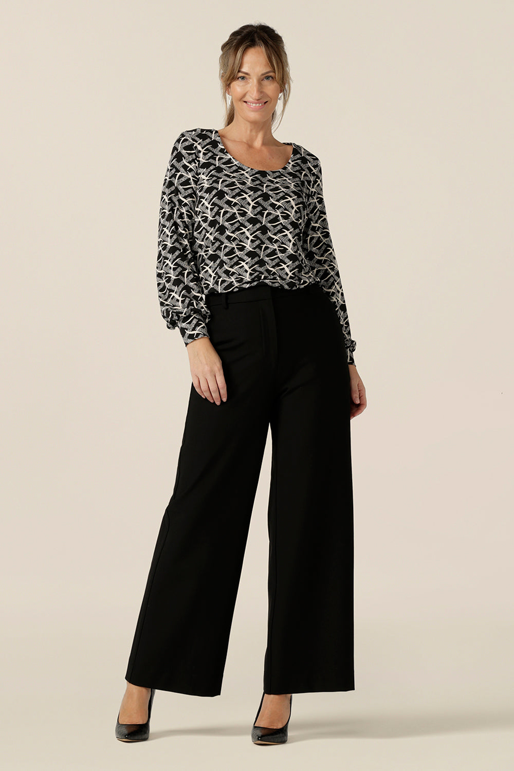 A size 10 woman wears a long sleeve top with bishop sleeve cuffs and a scoop neck in black and white print jersey. with tailored, wide leg black pants. Made in Australia by Australian and New Zealand women's clothing brand, L&F this women's work top is available to shop in sizes 8 to 24.