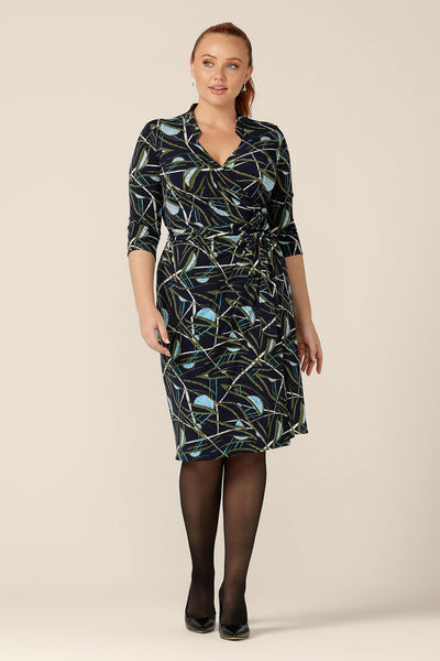 A size 12 woman wears an Australian-made jersey wrap dress by Australian and New Zealand women's clothing label, L&F. Featuring a blue, green and white abstract print on navy jersey, this wrap dress has 3/4 sleeves and faux mandarin collar detail. This wrap dress is available to shop in an inclusive size range of 8 to 24.