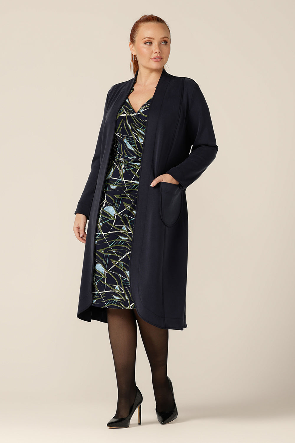 A size 12 woman wears the Marant Trenchcoat in Bluestone by Australian and New Zealand size inclusive clothing label, L&F. A collarless, open fronted coat with tie belt and patch pockets, this navy coat is worn with a jersey wrap dress for winter workwear layering.