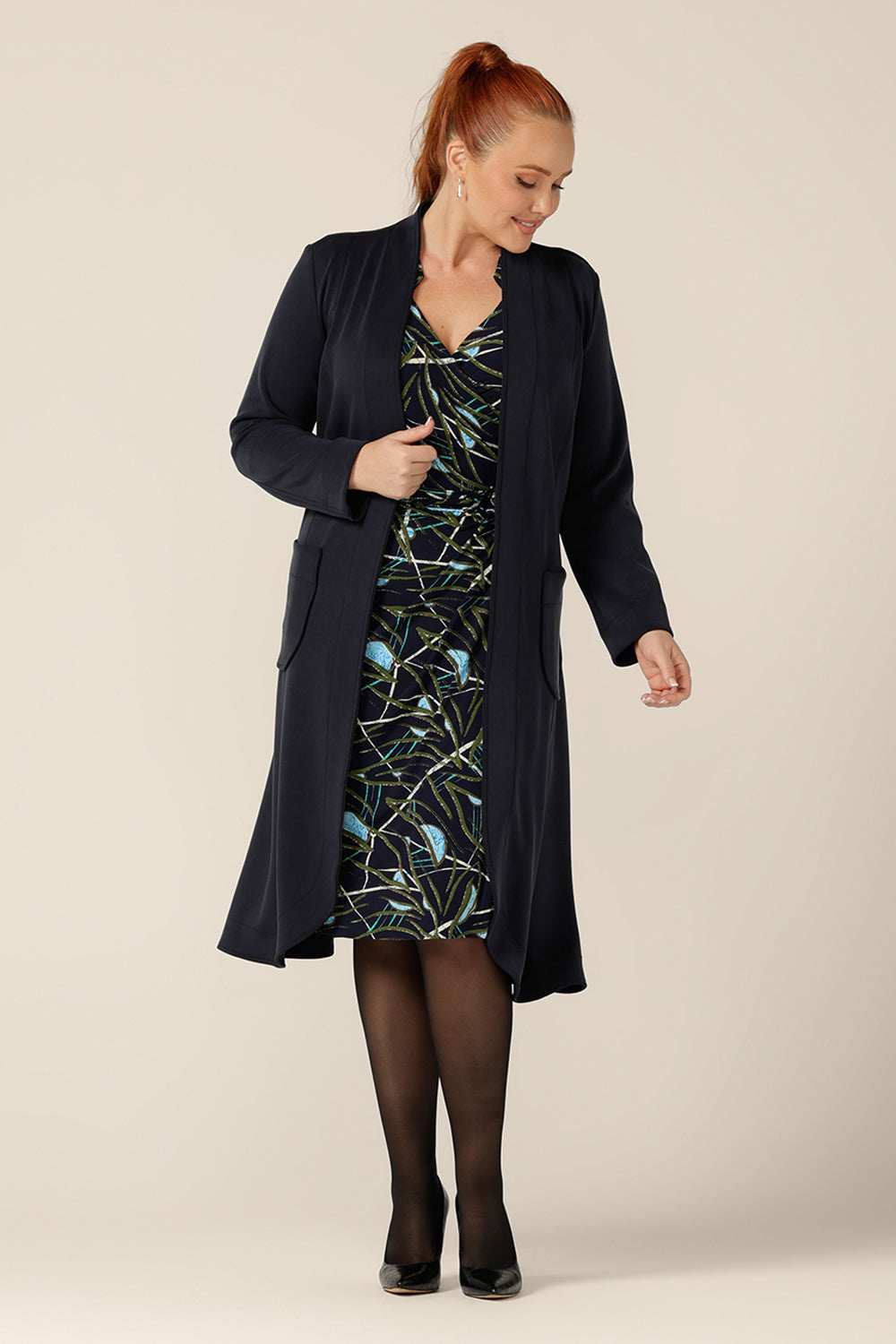 A size 12 woman wears the Marant Trenchcoat in Bluestone by Australian and New Zealand size inclusive clothing label, L&F. A collarless, open fronted coat with tie belt and patch pockets, this navy coat is worn with a jersey wrap dress, both of which are made in Australia.